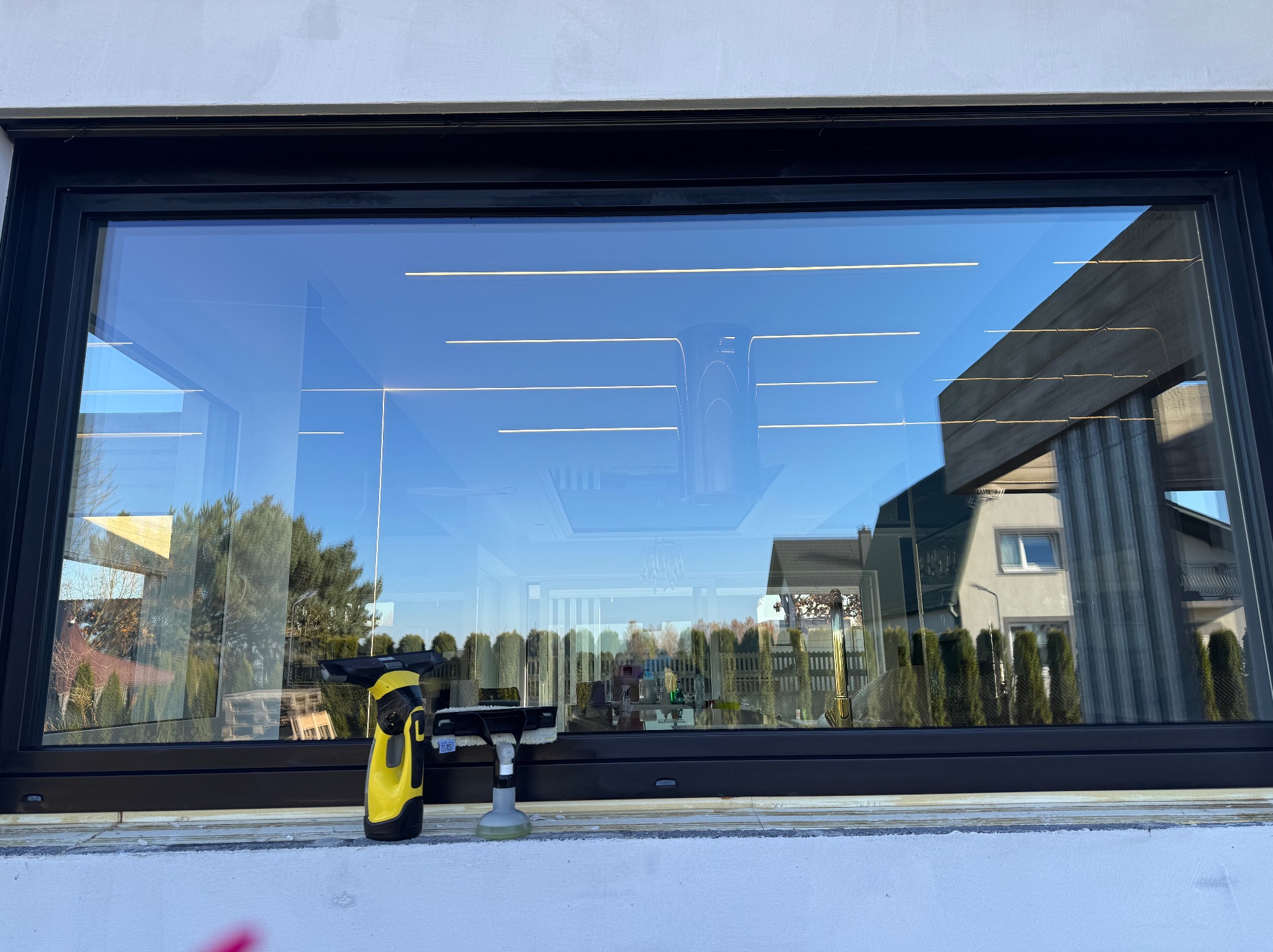Wyraźne odbicie w umytym oknie z czarną ramą. Na parapecie żółty odkurzacz do okien Kärcher i spryskiwacz. Widok na zewnątrz: domy, drzewa, błękitne niebo.