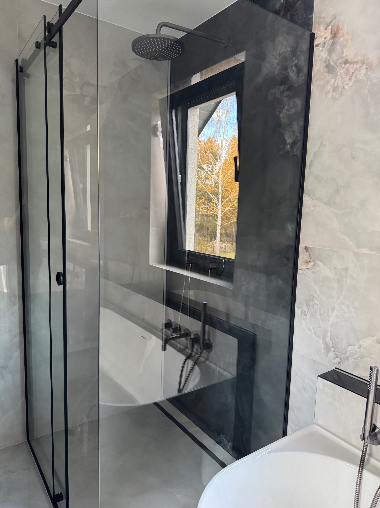 Nowoczesna kabina prysznicowa z czarnymi ramami i deszczownicą, widok na okno z jesiennym drzewem. Marmurowe płytki w odcieniach szarości i bieli. Minimalistyczny design.