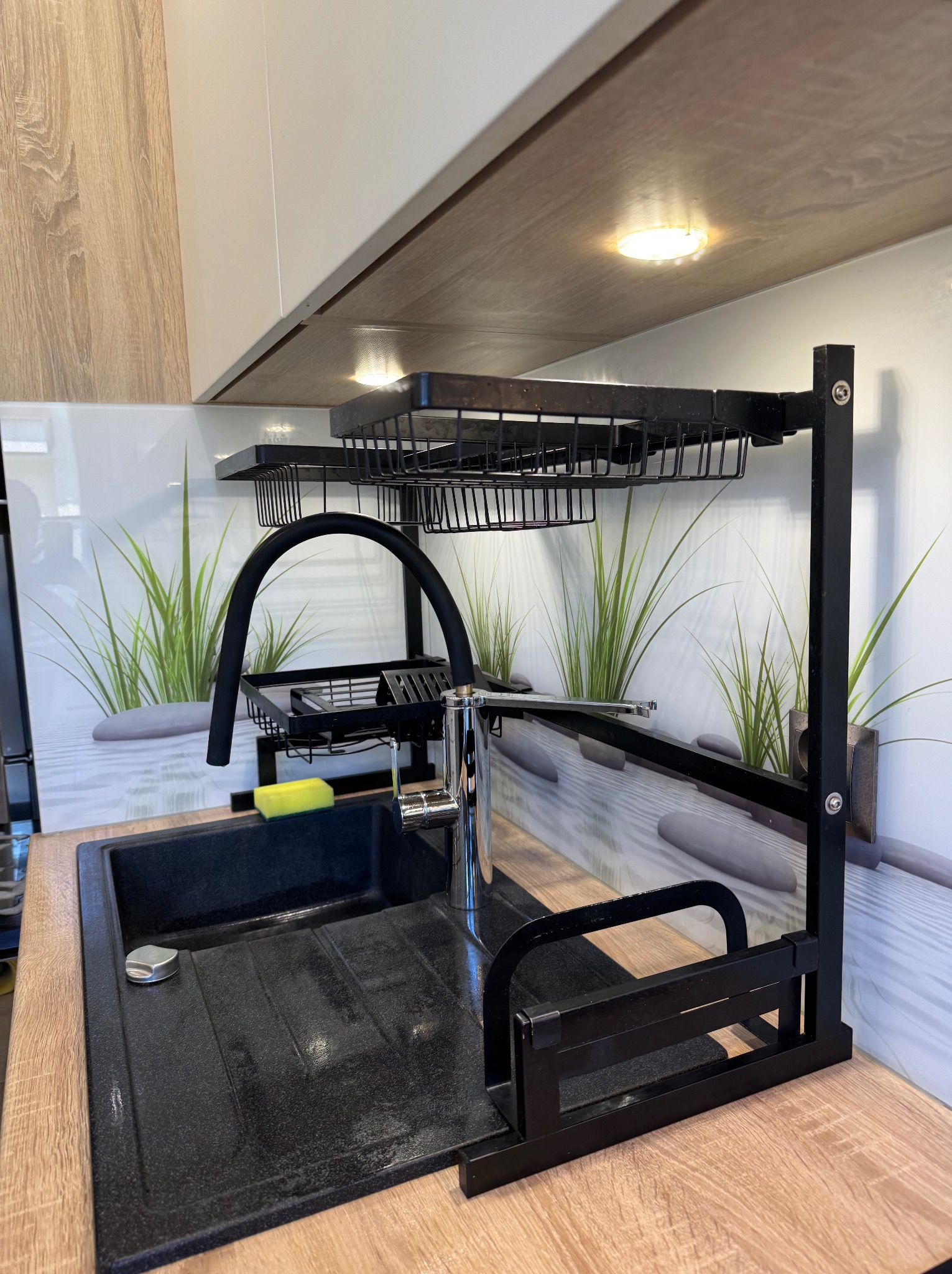 Czysty, czarny zlew granitowy z chromowaną baterią i ociekaczem na naczynia. Na blacie leży gąbka. Tło stanowi panel z motywem trawy i kamieni.