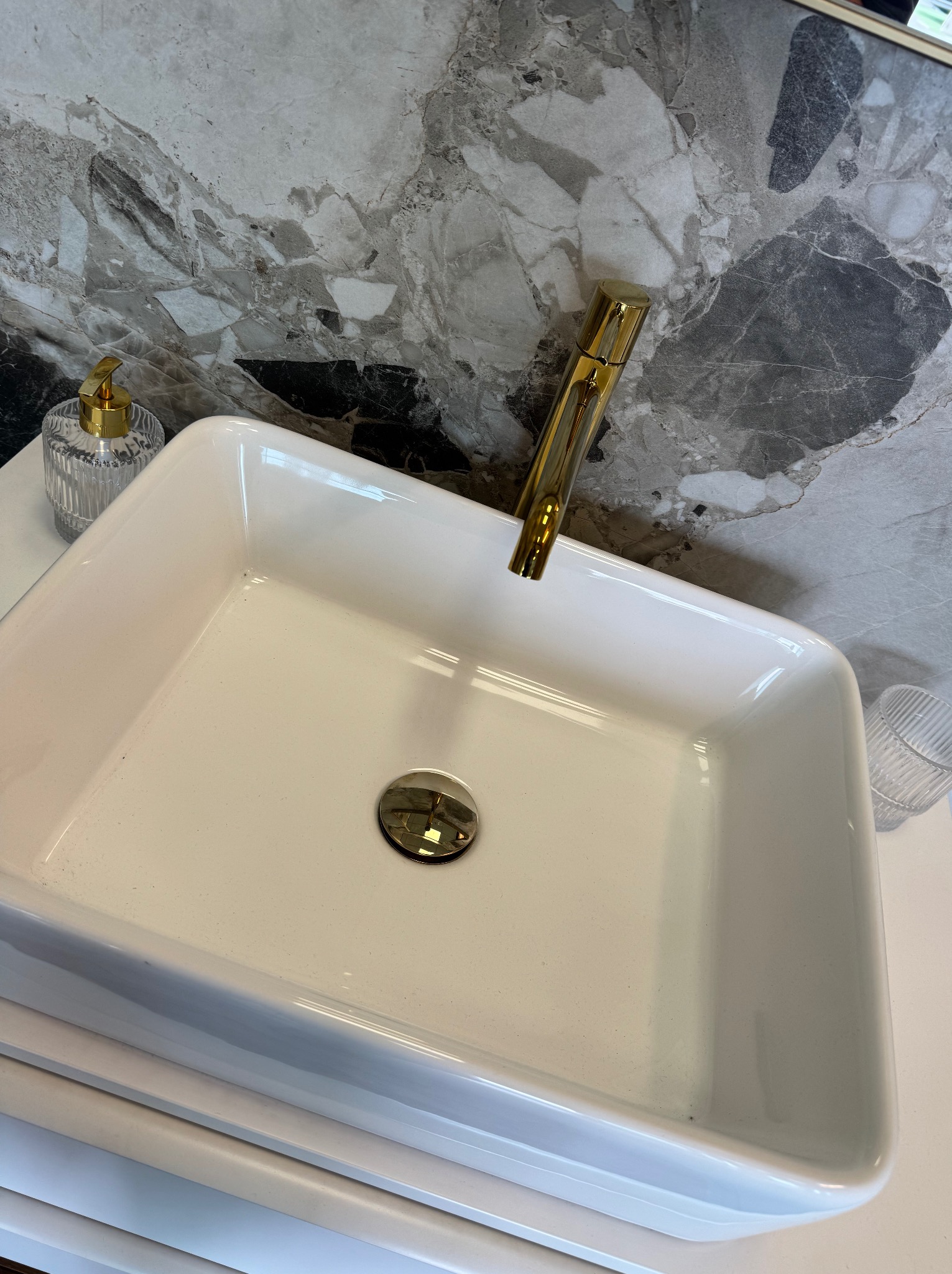 Biała, prostokątna umywalka z złotą armaturą na tle marmurowej ściany. Widoczny dozownik mydła i szklanka. Czystość i elegancja w detalu.