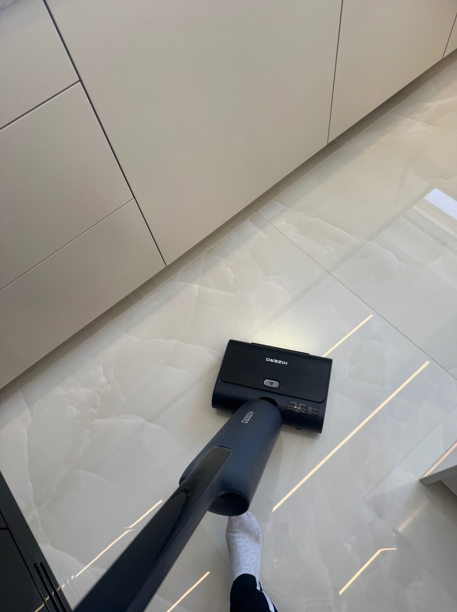 Błyszcząca, marmurowa podłoga czyszczona nowoczesnym mopem elektrycznym HiZero w jasnym wnętrzu kuchni z kremowymi szafkami. Widoczna stopa osoby obsługującej urządzenie.