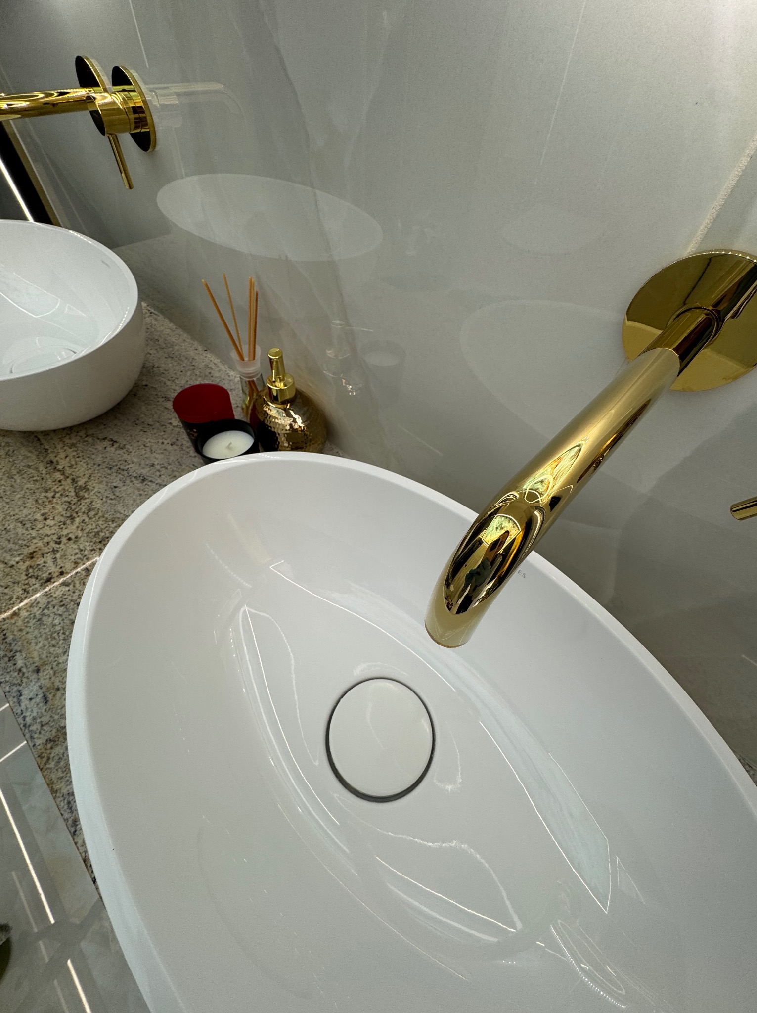 Biała, owalna umywalka nablatowa z złotą baterią w łazience. Obok dekoracje: świeca i dyfuzor zapachowy. Marmurowe ściany i blat. Ujęcie z góry, lekko obrócone.