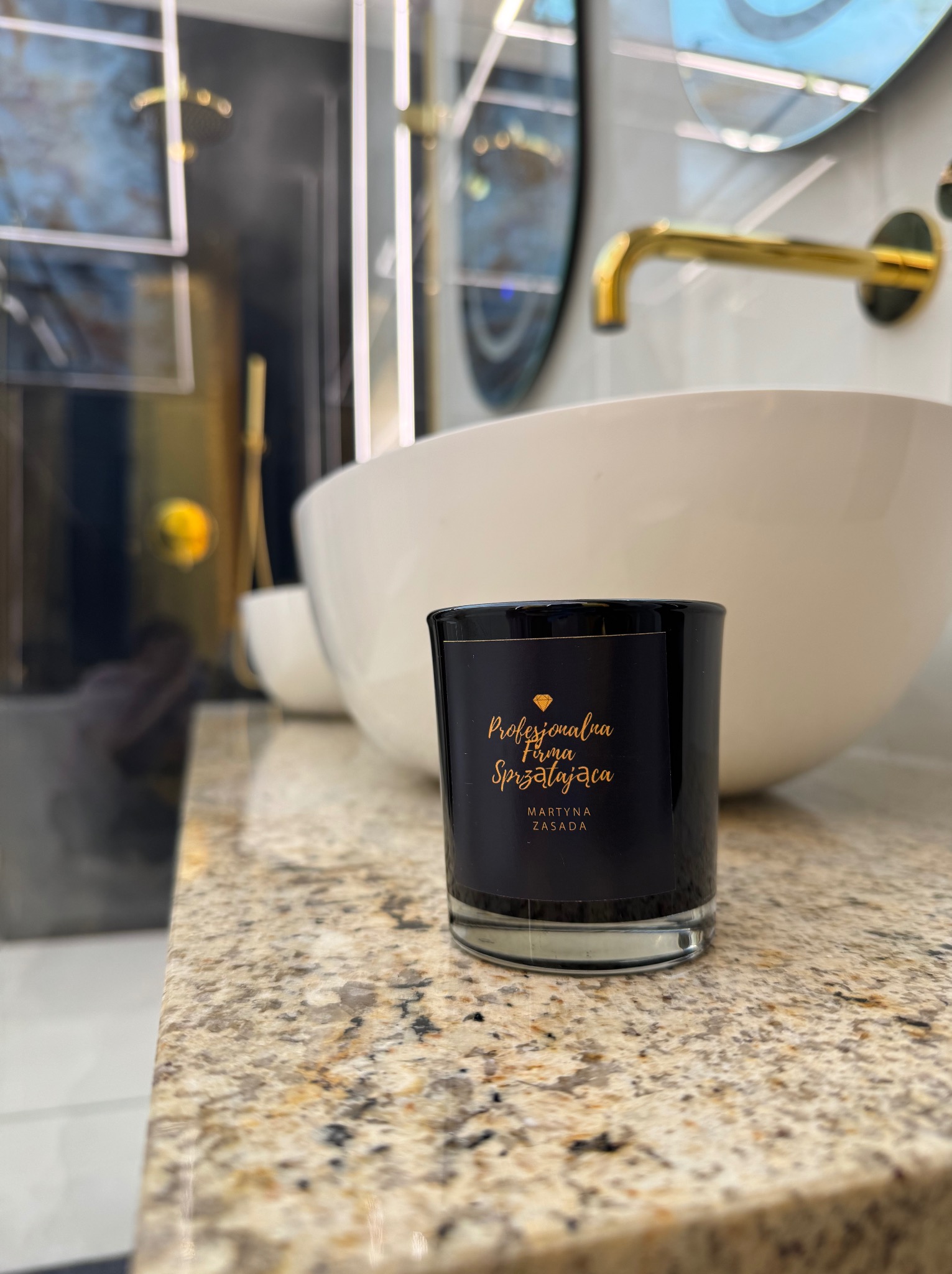 Elegancka łazienka z owalną umywalką i złotą armaturą. Na blacie stoi czarna świeca z napisem 'Profesjonalna Firma Sprzątająca'.