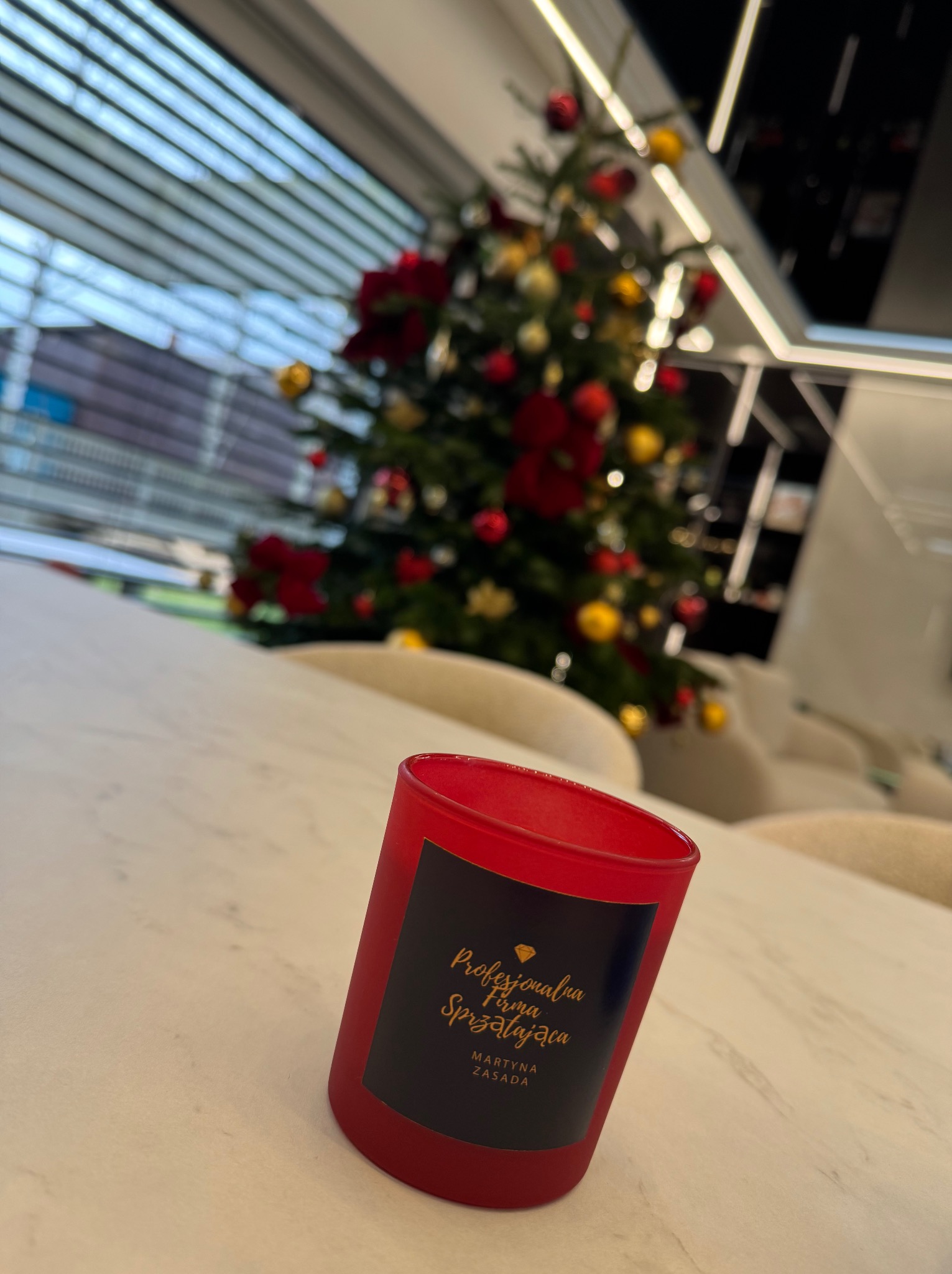 Czerwona świeca z logo firmy sprzątającej na stole, w tle choinka i jasne wnętrze biura. Świąteczny akcent w przestrzeni po sprzątaniu.
