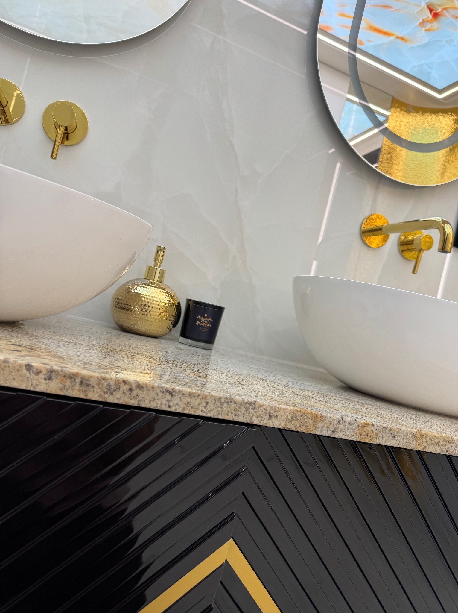 Białe umywalki nablatowe z złotą armaturą na blacie z kamienia. Czarna szafka z geometrycznym wzorem. Okrągłe lustra z podświetleniem. Luksusowe wykończenie łazienki.