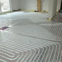 Białe rury systemu ogrzewania podłogowego ułożone spiralnie na podłodze pokrytej siatką, widoczne fragmenty czerwonych i niebieskich rur przy ścianach, żółta taśma zabezpieczająca wzdłuż ścian.