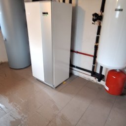 MULTI TECH TOMASZ MARCINIUK - Pompa ciepła w kotłowni: biała jednostka wewnętrzna, zbiornik buforowy i instalacja rurowa na ścianie. Widoczne przyłącza i manometry. Podłoga z płytek.