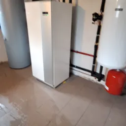 Pompa ciepła w kotłowni: biała jednostka wewnętrzna, zbiornik buforowy i instalacja rurowa na ścianie. Widoczne przyłącza i manometry. Podłoga z płytek.