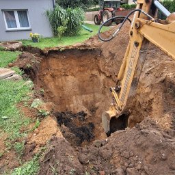 MULTI TECH TOMASZ MARCINIUK - Żółta koparka CASE pogłębia wykop w ziemi, obok traktor i domy jednorodzinne. Widoczne warstwy gleby. Prace ziemne w toku.