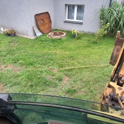 MULTI TECH TOMASZ MARCINIUK - Widok z kabiny koparki na trawnik z elementami wykopu pod szambo, w tle budynek z oknami. Perspektywa operatora maszyny budowlanej.