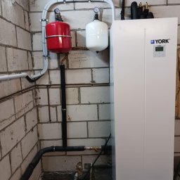 MULTI TECH TOMASZ MARCINIUK - Biała pompa ciepła York z instalacją hydrauliczną (czerwony i biały zbiornik) przy ścianie z bloczków. Widoczne rury i manometry.