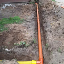 Pomarańczowe rury kanalizacyjne ułożone w wykopie ziemnym wzdłuż budynku, z widoczną żółtą rurą karbowaną. W tle łopata wbita w ziemię.