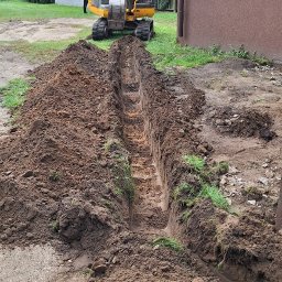 MULTI TECH TOMASZ MARCINIUK - Żółta koparka wykonuje wykop liniowy w ziemi, widoczny zarys fundamentów budynku w tle. Prace ziemne przy użyciu ciężkiego sprzętu.