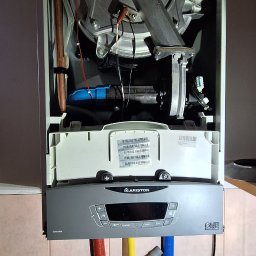 MULTI TECH TOMASZ MARCINIUK - Otwarty piec gazowy Ariston Clas One z widocznymi podłączeniami hydraulicznymi (czerwony, żółty, niebieski) na tle kafelkowanej ściany.