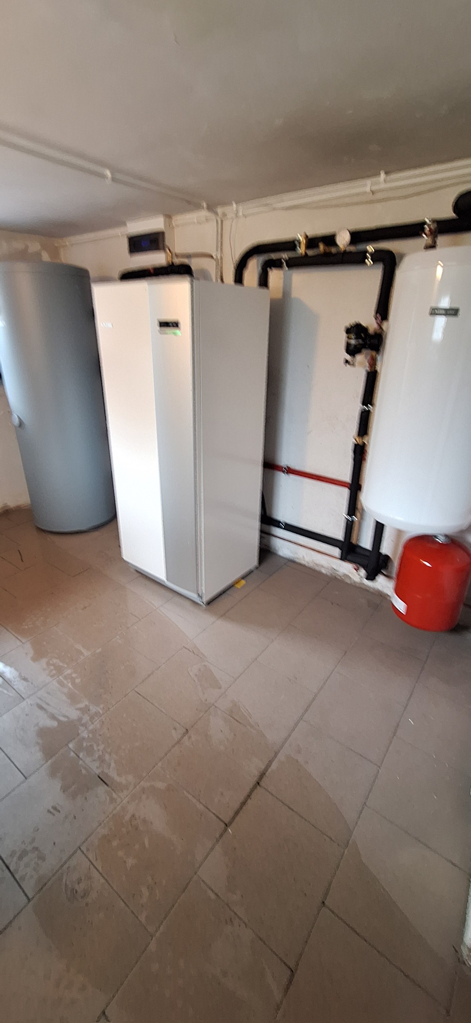 Pompa ciepła w kotłowni: biała jednostka wewnętrzna, zbiornik buforowy i instalacja rurowa na ścianie. Widoczne przyłącza i manometry. Podłoga z płytek.