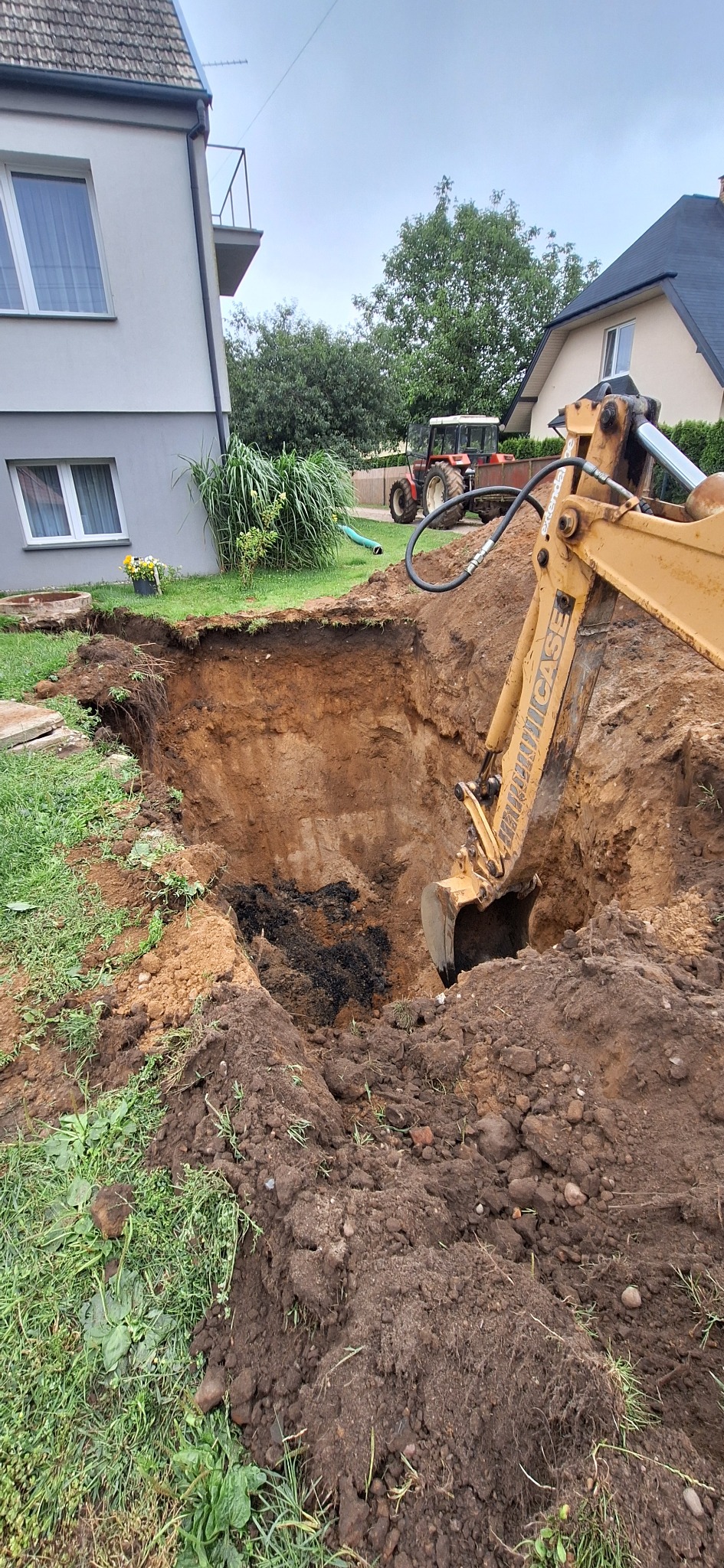 Żółta koparka CASE pogłębia wykop w ziemi, obok traktor i domy jednorodzinne. Widoczne warstwy gleby. Prace ziemne w toku.