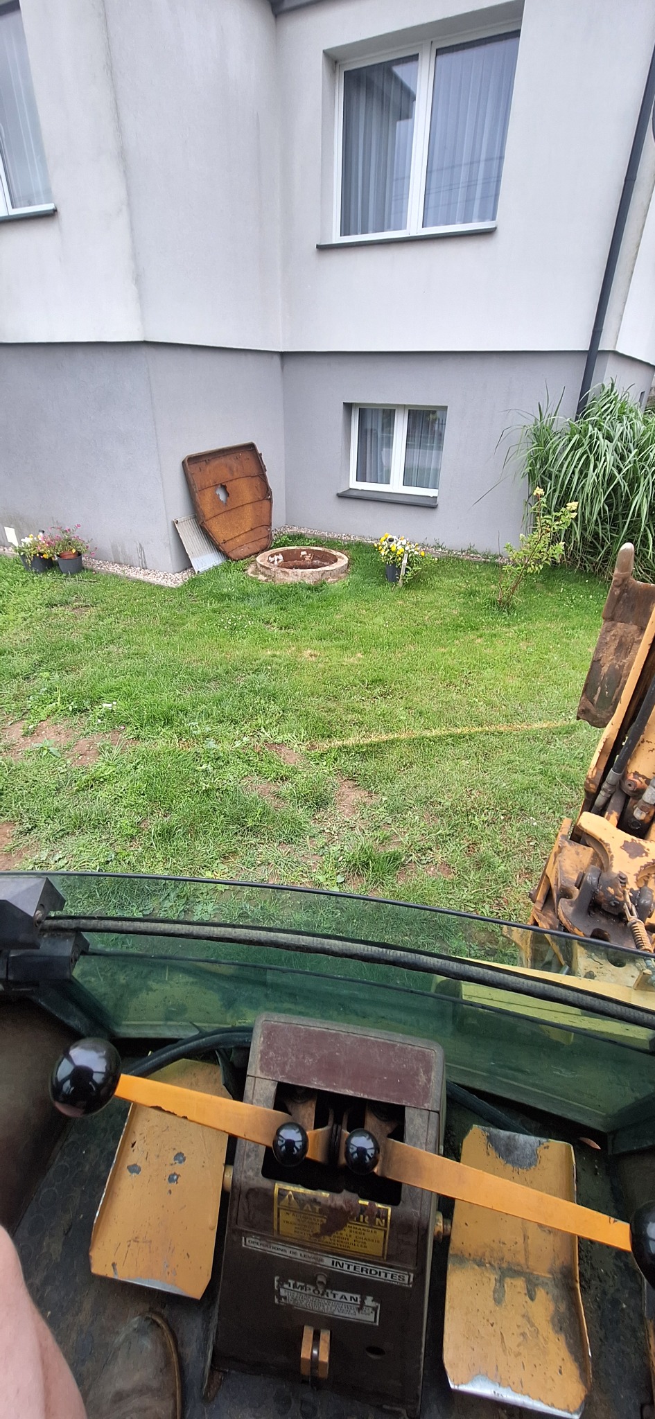 Widok z kabiny koparki na trawnik z elementami wykopu pod szambo, w tle budynek z oknami. Perspektywa operatora maszyny budowlanej.