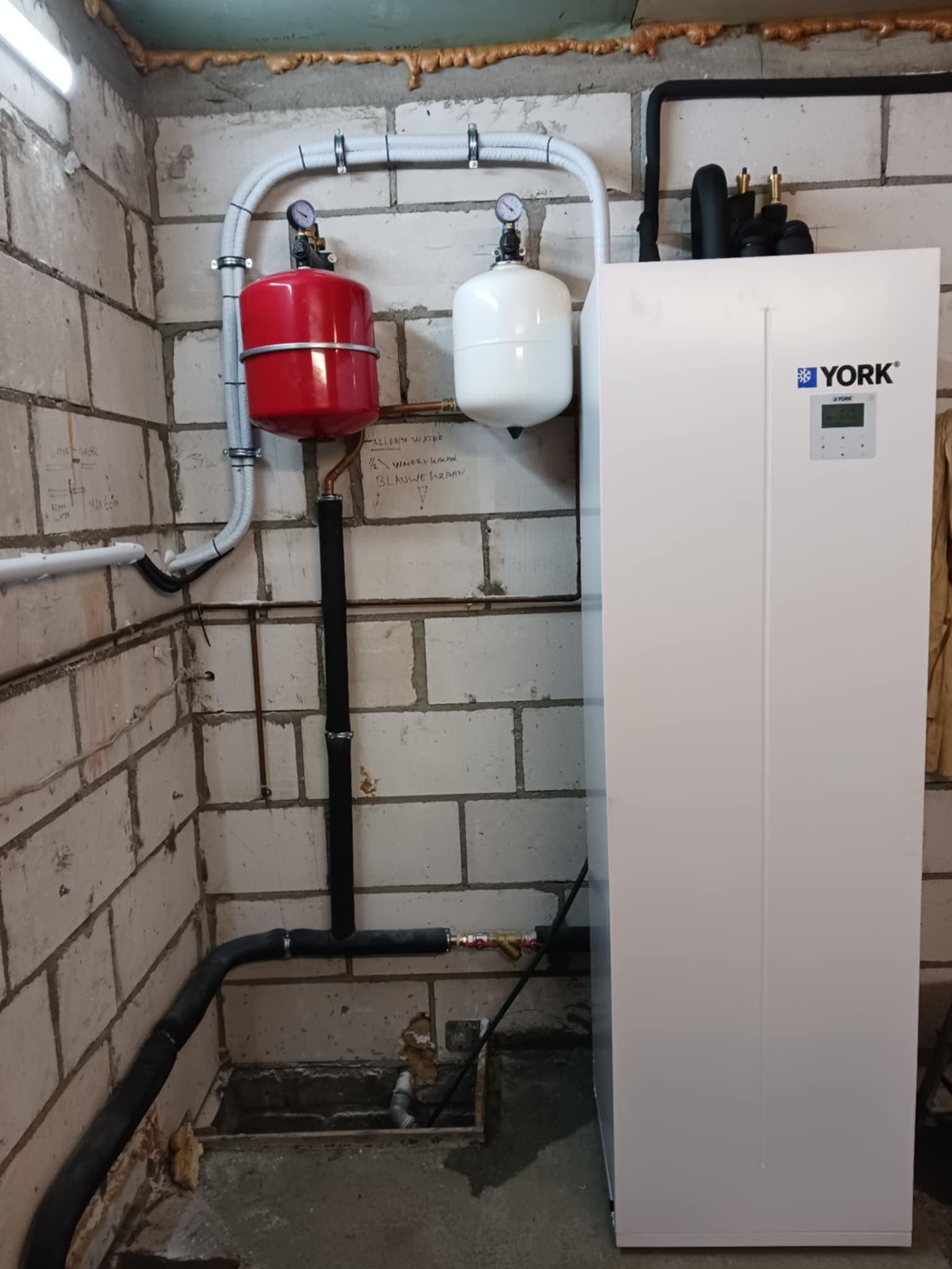 Biała pompa ciepła York z instalacją hydrauliczną (czerwony i biały zbiornik) przy ścianie z bloczków. Widoczne rury i manometry.