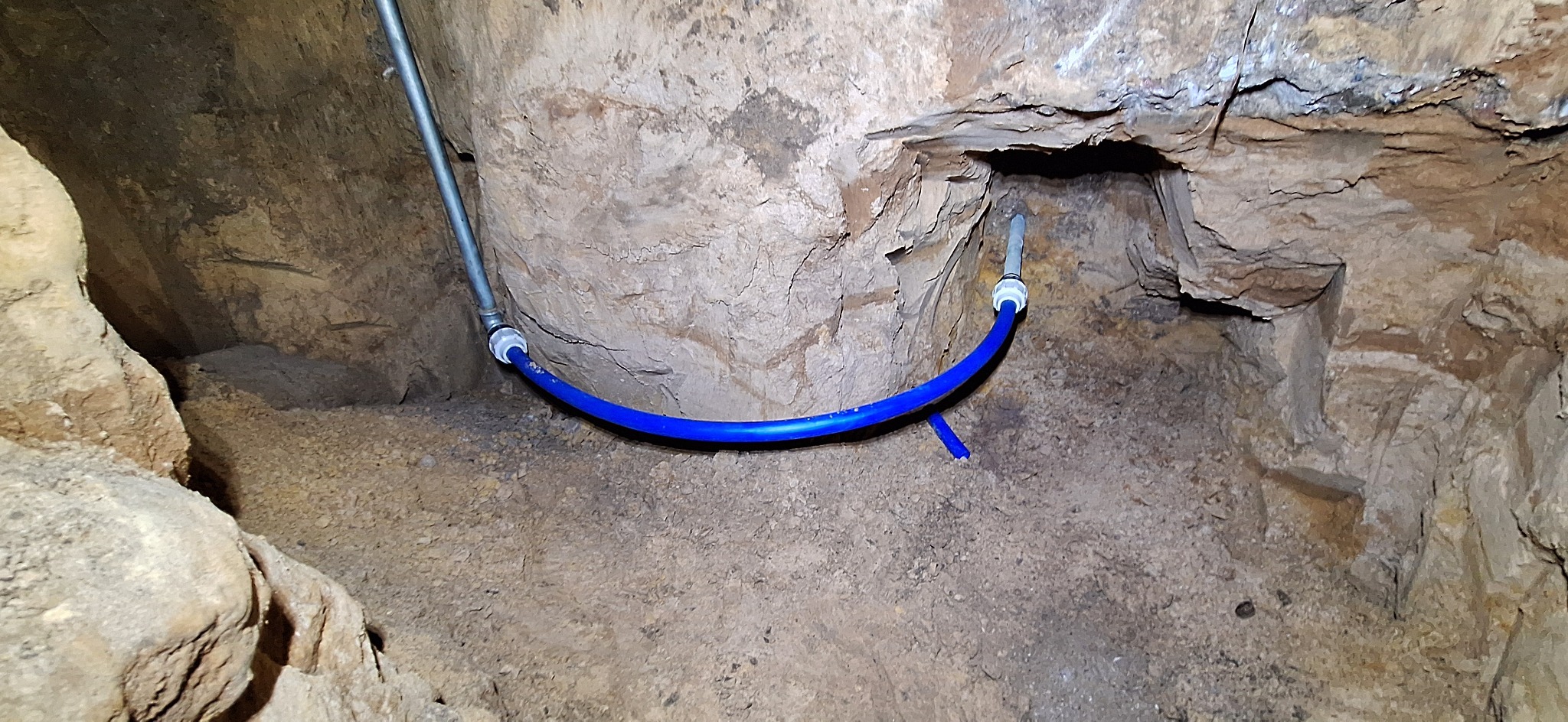 Niebieska rura wodna z łącznikami, poprowadzona w wykopie o nierównych ścianach z ziemi i kamieni. Szara rura pionowa w tle.