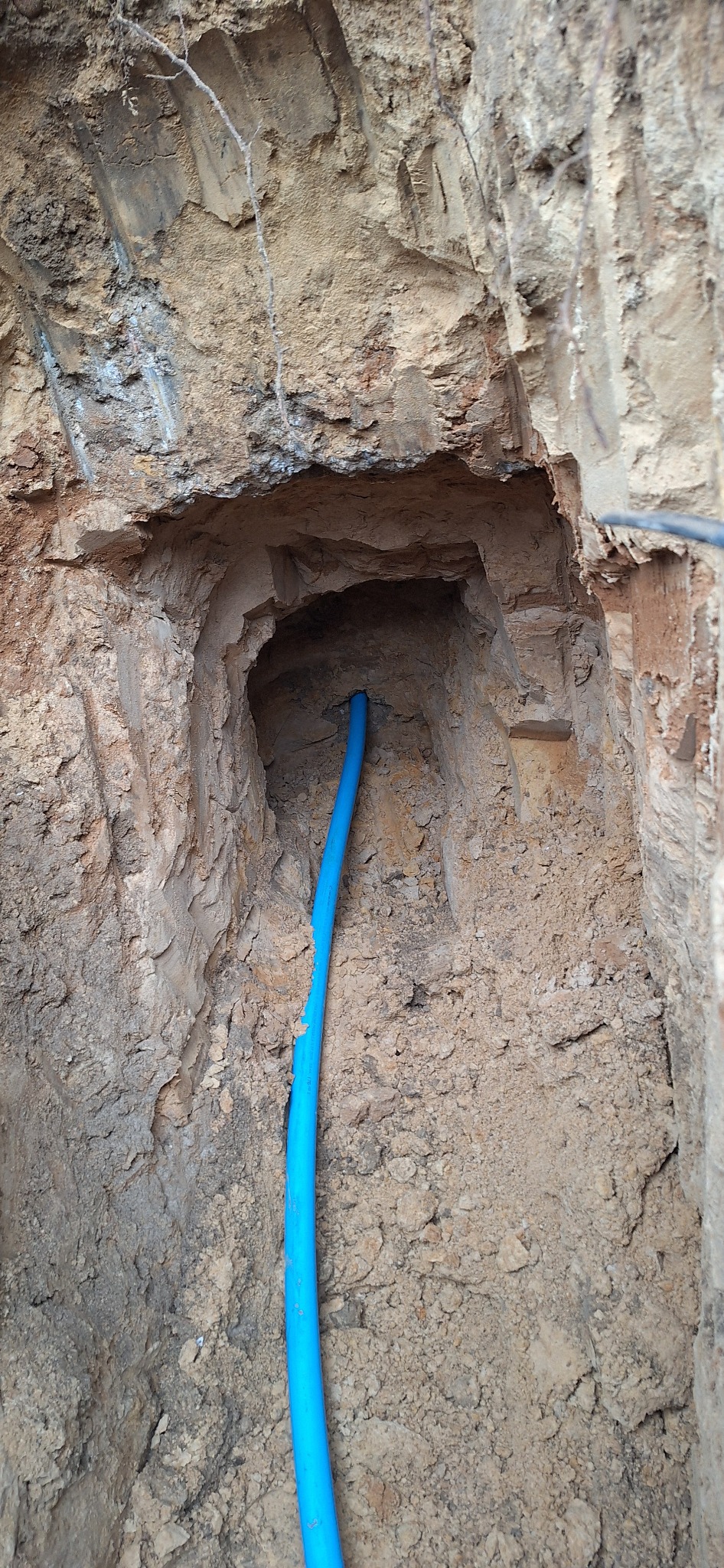 Niebieska rura w wykopie o nierównych ścianach z piasku i gliny. W tle widoczne korzenie i fragmenty kabli. Ujęcie z bliska, pionowe.