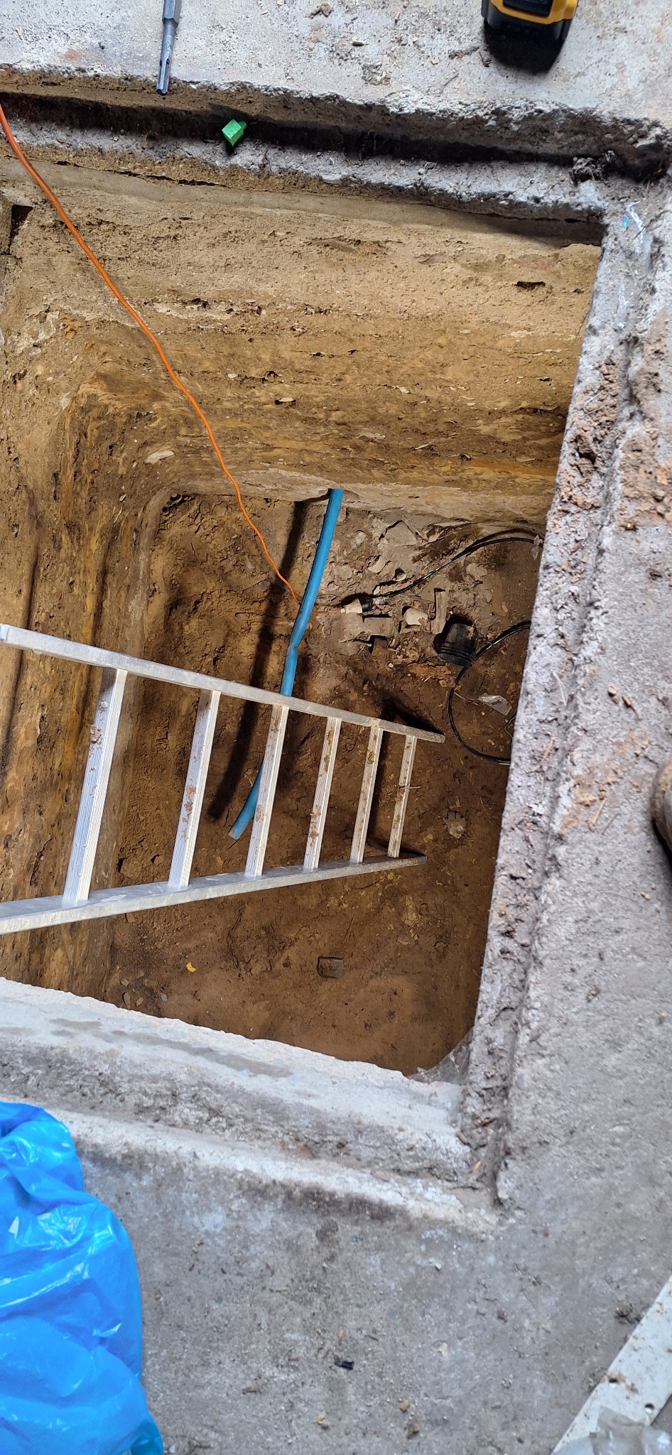 Wykop z drabiną i niebieską rurą. Widoczne narzędzia na górze. Pomarańczowy kabel biegnie w dół wykopu. Prace ziemne lub instalacyjne w toku.