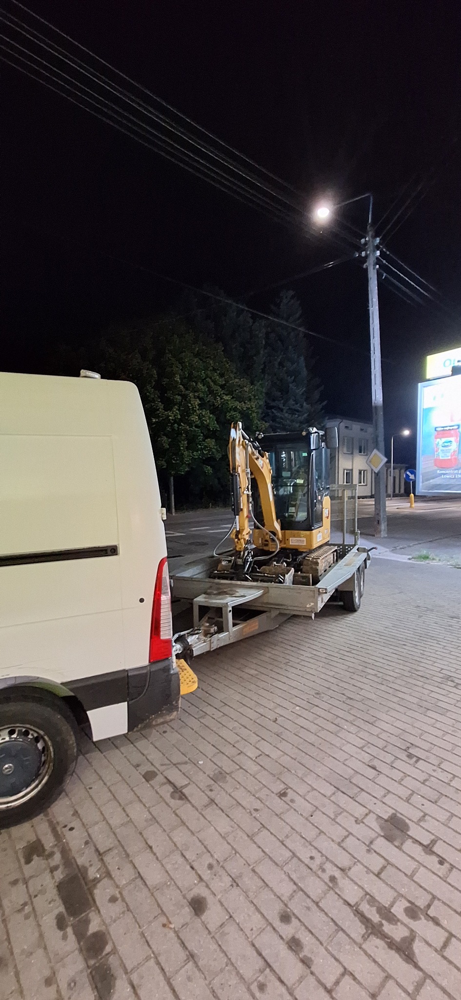 Żółta minikoparka na przyczepie, transportowana białym busem na tle nocnego krajobrazu miejskiego. Widoczny słup oświetleniowy i fragment reklamy.