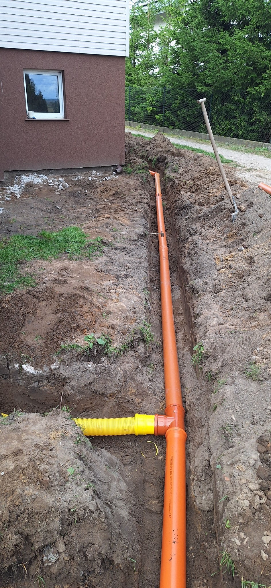 Pomarańczowe rury kanalizacyjne ułożone w wykopie ziemnym wzdłuż budynku, z widoczną żółtą rurą karbowaną. W tle łopata wbita w ziemię.