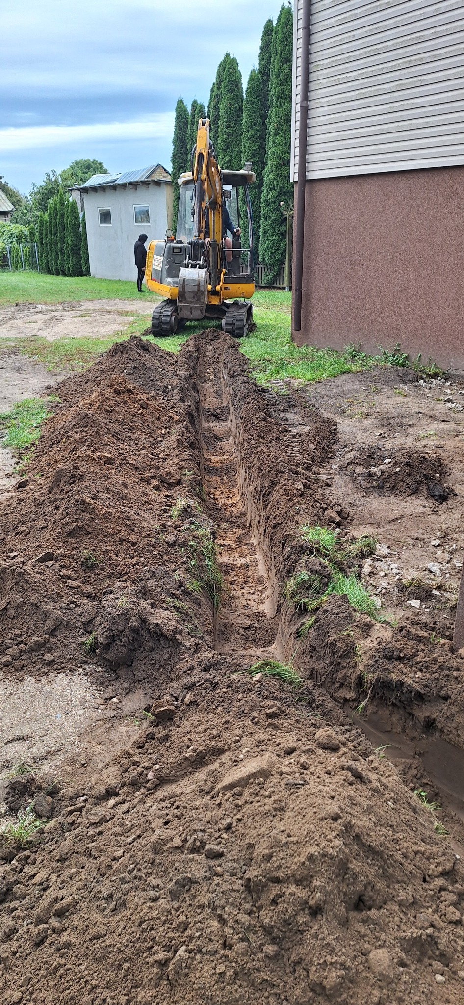 Żółta koparka wykonuje wykop liniowy w ziemi, widoczny zarys fundamentów budynku w tle. Prace ziemne przy użyciu ciężkiego sprzętu.