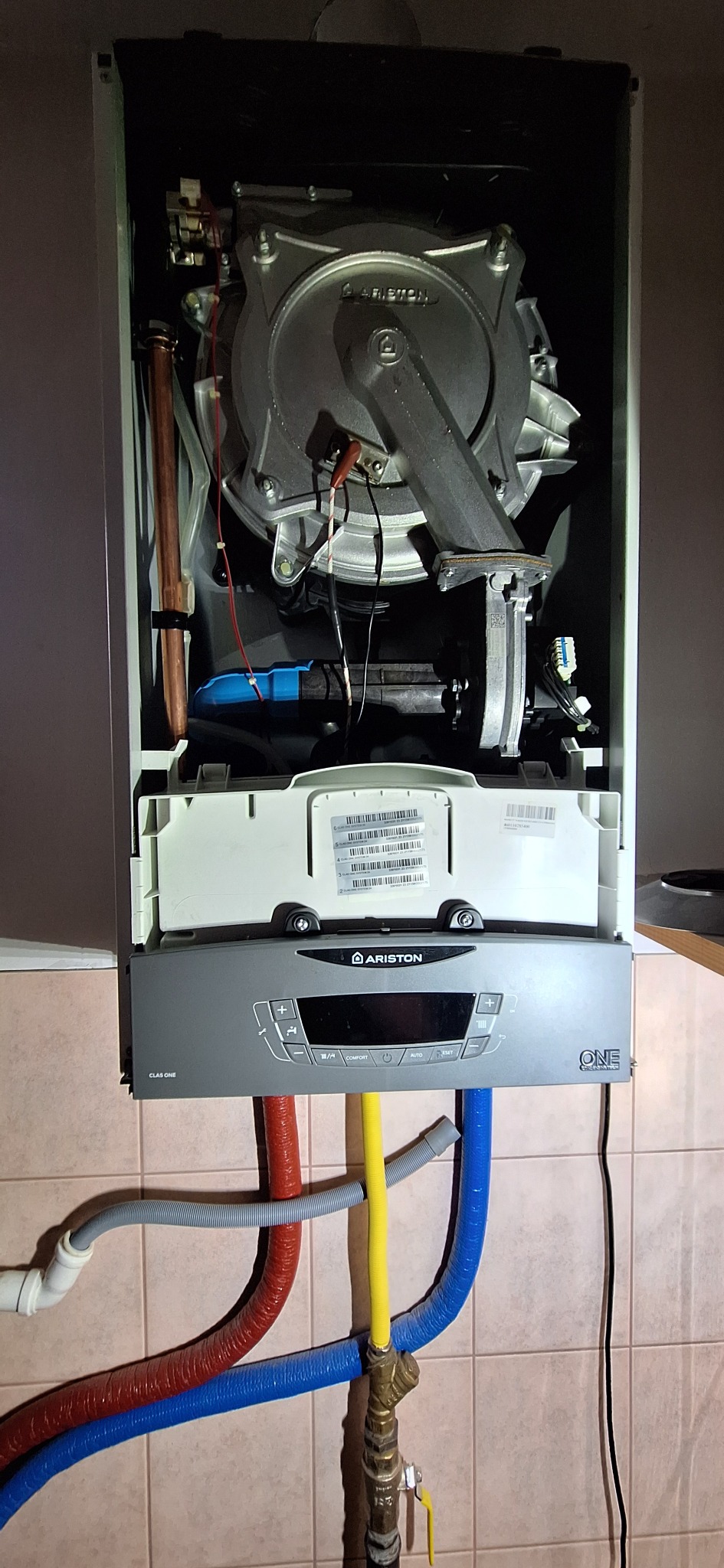 Otwarty piec gazowy Ariston Clas One z widocznymi podłączeniami hydraulicznymi (czerwony, żółty, niebieski) na tle kafelkowanej ściany.