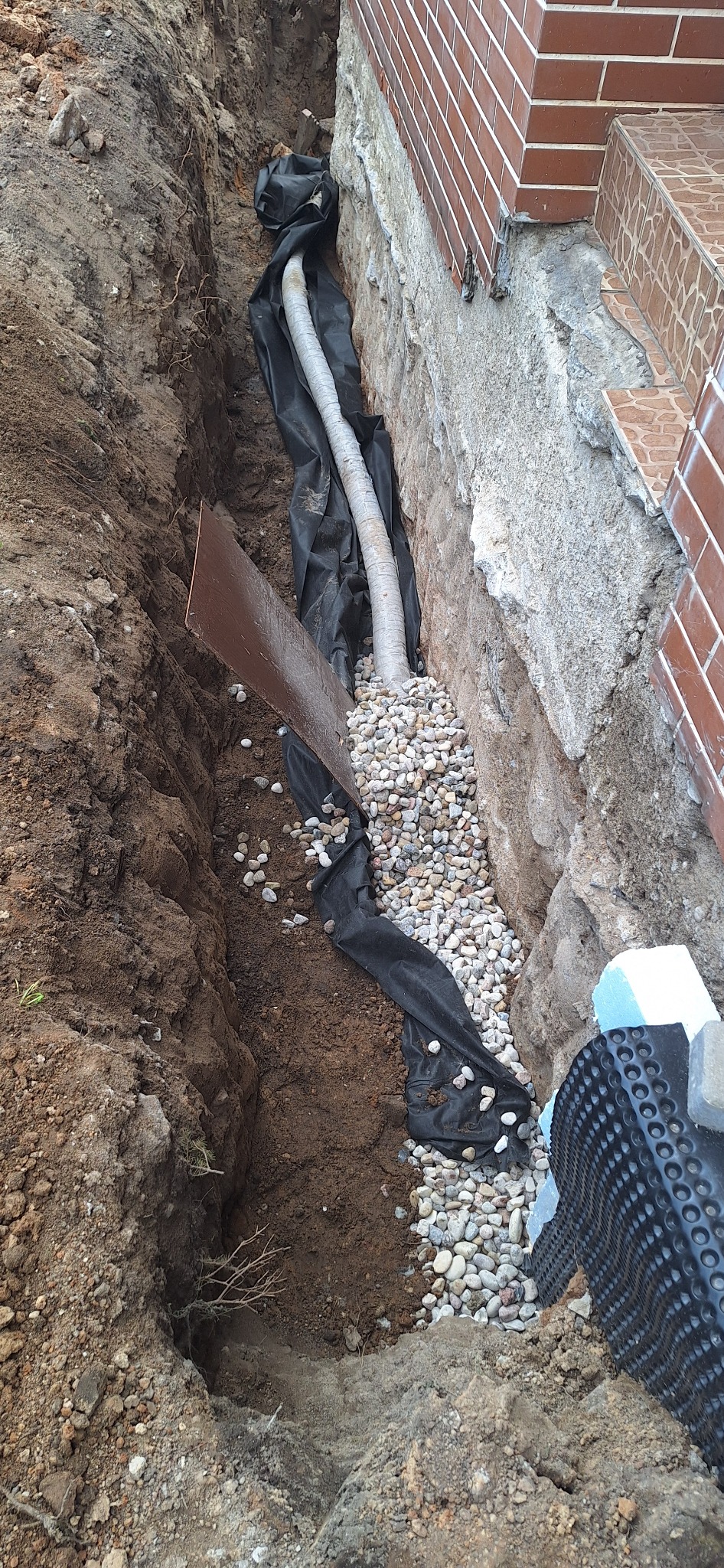 Wykop fundamentu budynku z ułożoną rurą drenażową w geowłókninie, obsypaną kamieniami. Widoczna ściana fundamentowa i fragment schodów wejściowych.