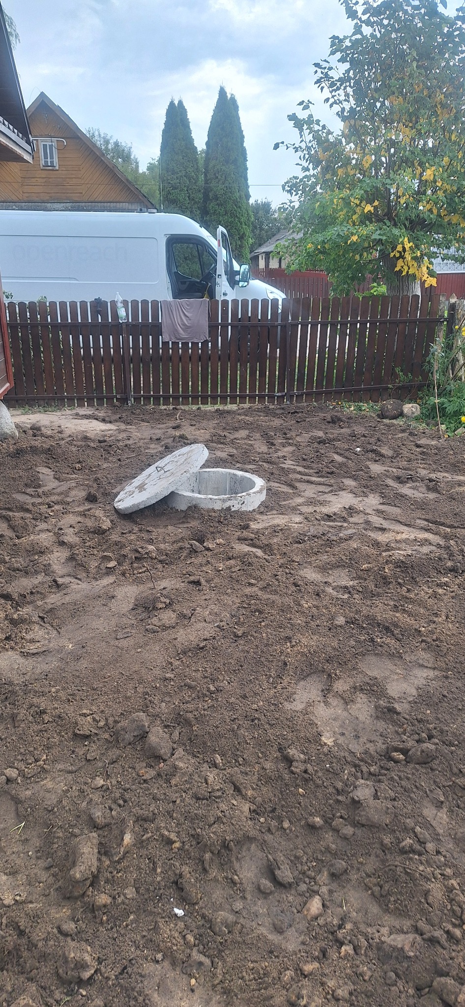 Otwarty betonowy krąg studzienny na ziemi, za nim brązowe ogrodzenie i biały samochód dostawczy. Teren przygotowany pod dalsze prace.