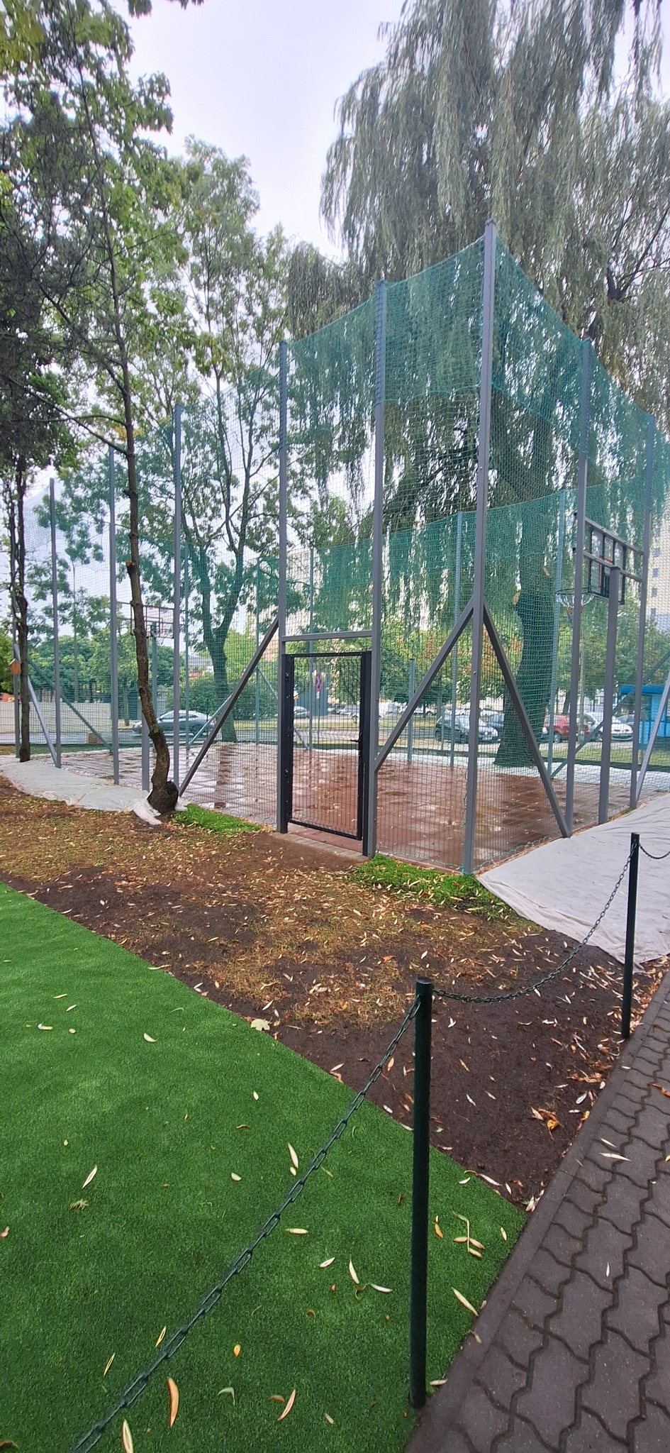 Nowo wybudowane boisko sportowe z zieloną sztuczną trawą, siatką ochronną i metalową bramką wejściową. Widoczny fragment chodnika i drzewa w tle.