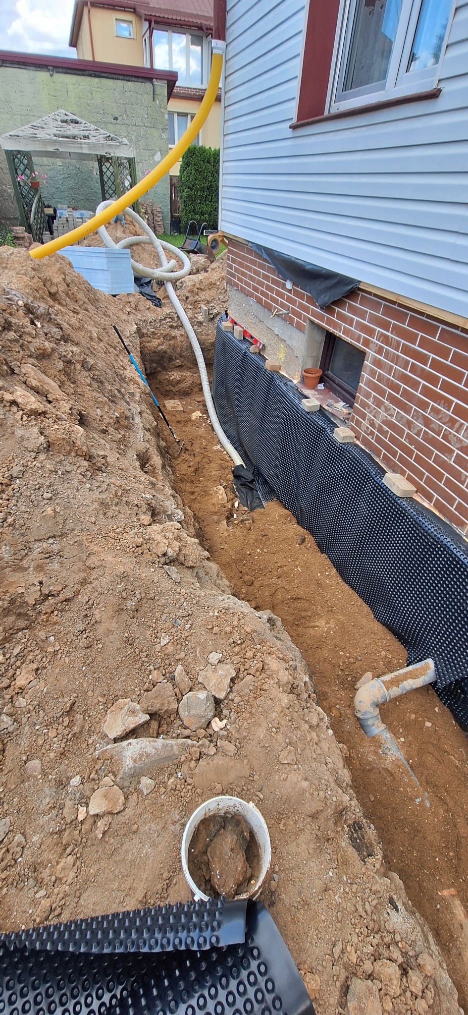 Wykopany rów wzdłuż fundamentów domu z zabezpieczeniem w postaci folii kubełkowej. Widoczne rury odprowadzające wodę i narzędzie w wykopie. Prace ziemne.