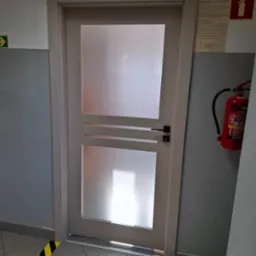 Białe drzwi wewnętrzne z matowym przeszkleniem i poziomymi listwami dekoracyjnymi, czarna klamka i klucz w zamku, widok na ścianę z gaśnicą i instrukcją przeciwpożarową.