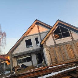 R&S DOORS SPÓŁKA Z OGRANICZONĄ ODPOWIEDZIALNOŚCIĄ - Dom w stanie surowym otwartym z zamontowanymi oknami aluminiowymi o nietypowym kształcie, drewniane deski zabezpieczające otwór, widoczne białe bloczki konstrukcyjne i przygotowane drewno na dachu.