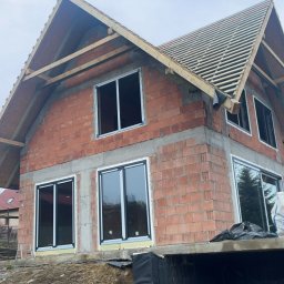 R&S DOORS SPÓŁKA Z OGRANICZONĄ ODPOWIEDZIALNOŚCIĄ - Surowy stan budynku jednorodzinnego z zamontowanymi oknami z czarnymi ramami, widoczne cegły, szare wykończenia, nieotynkowana elewacja, konstrukcja dachu w trakcie budowy, pomarańczowa rura...