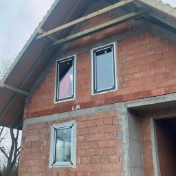 R&S DOORS SPÓŁKA Z OGRANICZONĄ ODPOWIEDZIALNOŚCIĄ - Surowy stan budynku w trakcie budowy z trzema oknami z czarnymi ramami na tle czerwonej cegły, widoczne drewniane elementy konstrukcji dachu i czarna izolacja fundamentów.