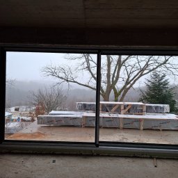 R&S DOORS SPÓŁKA Z OGRANICZONĄ ODPOWIEDZIALNOŚCIĄ - Duże przesuwne okno w surowym stanie, widok na zimowy krajobraz z drzewami i paletami z materiałami budowlanymi na zewnątrz.