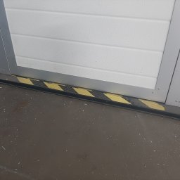 R&S DOORS SPÓŁKA Z OGRANICZONĄ ODPOWIEDZIALNOŚCIĄ - Fragment segmentowej bramy garażowej z uszczelką dolną z czarno-żółtymi pasami ostrzegawczymi, widok z bliska na posadzkę z drobnymi zabrudzeniami.