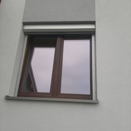 R&S DOORS SPÓŁKA Z OGRANICZONĄ ODPOWIEDZIALNOŚCIĄ - Brązowe okno z częściowo opuszczoną szarą roletą zewnętrzną na tle szarej, teksturowanej elewacji budynku.