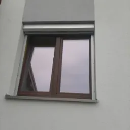 Brązowe okno z częściowo opuszczoną szarą roletą zewnętrzną na tle szarej, teksturowanej elewacji budynku.