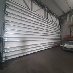 R&S DOORS SPÓŁKA Z OGRANICZONĄ ODPOWIEDZIALNOŚCIĄ - Szeroka segmentowa brama garażowa wewnątrz hali magazynowej, widoczne prowadnice i mechanizm podnoszenia, metalowa konstrukcja budynku w tle.