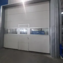 Biała segmentowa brama garażowa z oknami i małym drzwiami wejściowymi, widok z wnętrza hali, widoczny wózek transportowy i regały magazynowe w tle.