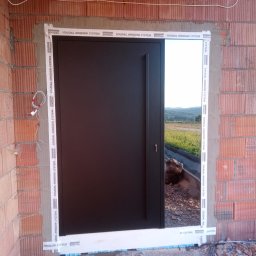 R&S DOORS SPÓŁKA Z OGRANICZONĄ ODPOWIEDZIALNOŚCIĄ - Montaż czarnych drzwi zewnętrznych w surowym murze z cegły, widoczne uszczelnienia Soudal Window System, otwarte drzwi ukazują krajobraz za domem.