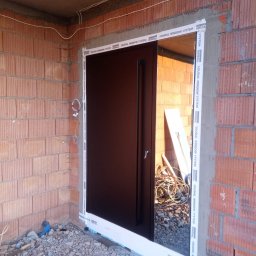 R&S DOORS SPÓŁKA Z OGRANICZONĄ ODPOWIEDZIALNOŚCIĄ - Montaż czarnych drzwi wejściowych w surowym stanie, widoczne cegły, przewody i materiały budowlane.