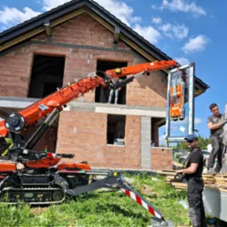 Montaż okna za pomocą minikoparki z przyssawkami na budowie domu z czerwonej cegły. Operator steruje maszyną, drugi pracownik pomaga w montażu.