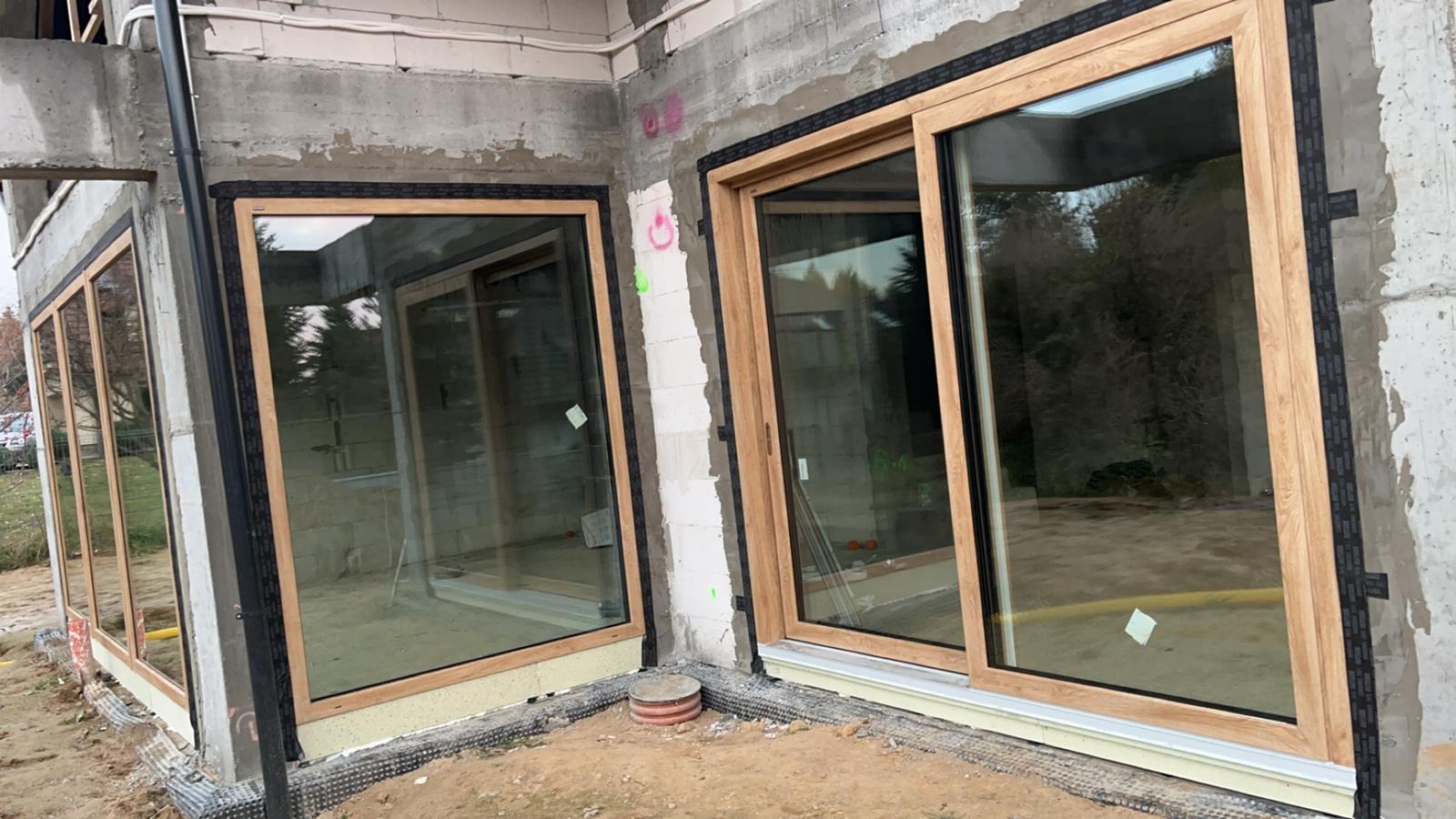 Montaż okien przesuwnych PCV o drewnopodobnej okleinie w surowym stanie budynku, widoczne pianka montażowa i taśmy uszczelniające.