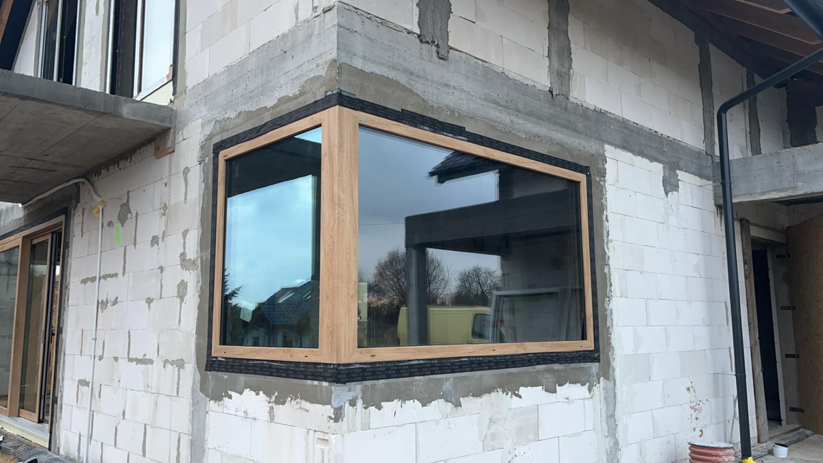 Narożne okno z drewnianą ramą zamontowane w surowej ścianie z betonu komórkowego, widoczne czarne uszczelnienie i odbicie otoczenia w szybie.