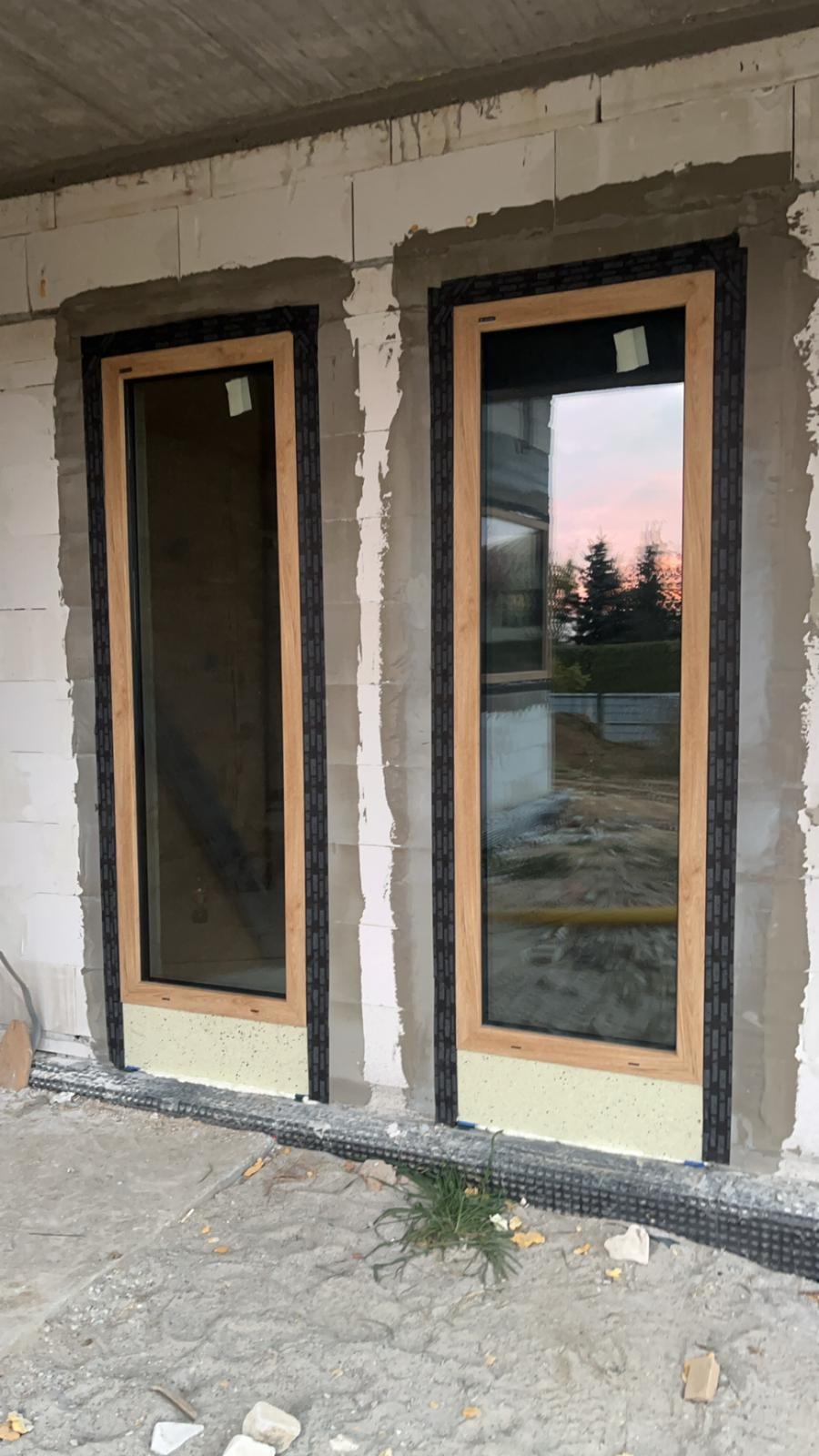 Dwa okna z drewnopodobną ramą, zamontowane w surowym murze z betonu komórkowego, widoczne zabezpieczenia montażowe i pianka izolacyjna u dołu, w szybach odbija się zachodzące słońce i drzewa.