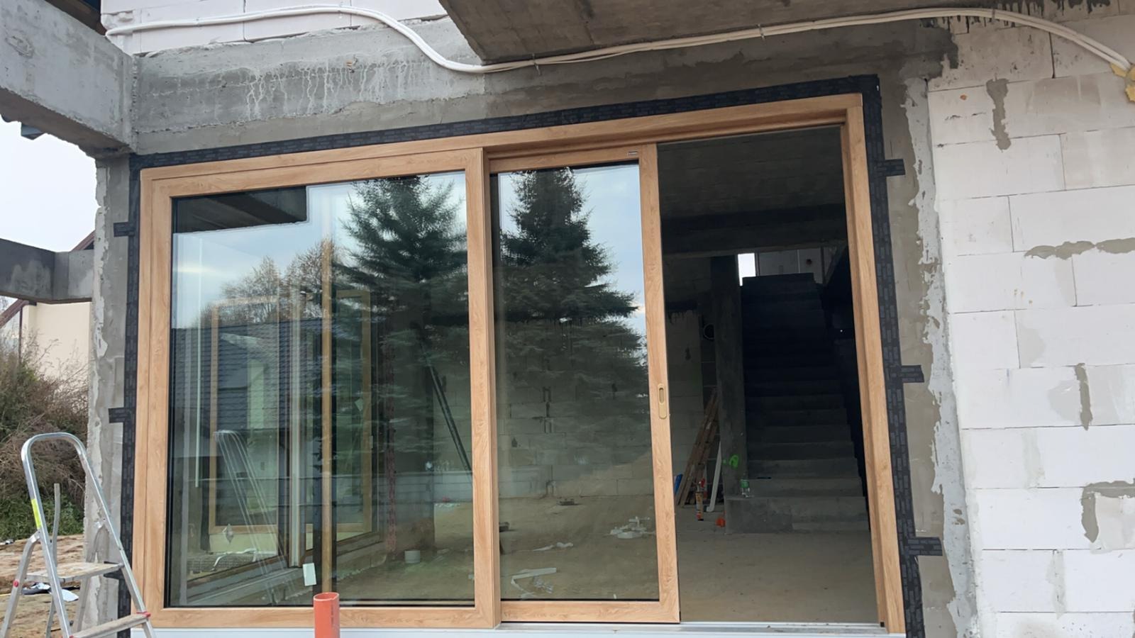 Montaż okna przesuwnego z jasną ramą na surowej ścianie z betonu komórkowego, widoczne schody wewnątrz budynku, drabina oparta o ścianę na zewnątrz.